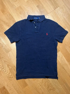Marinblå pikétröja Polo Ralph Lauren - Säljer denna klassisk och mycket stilrena marinblå pikétröja från Polo Ralph Lauren i storlek M slim fit med röd broderad logga på bröstet. Pikétröjn har korta ärmar, krage och knappar vid halsen. Perfekt till jeans eller shorts för en clean och stilren look. Vid frågor, extra bilder eller liknande är det bara att kontakta mig!