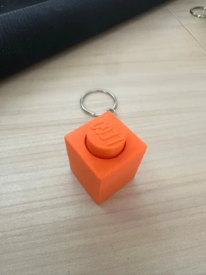 Orange LEGO-nyckelring kub clicker - Säljer en orange nyckelring formad som en stor LEGO-kloss. Tillverkad i plast och har en metallring för att fästa nycklar. Perfekt för dig som gillar LEGO eller vill ha en unik accessoar till väskan eller nyckelknippan.