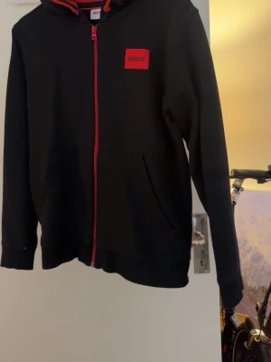 Svart hoodie med röd dragkedja från Hugo - Snygg svart hoodie från Hugo med röd dragkedja och röd logotyp på bröstet. Klassisk huva och fickor framtill. Perfekt för en stilren och avslappnad look. Tillverkad i mjukt material för extra komfort.