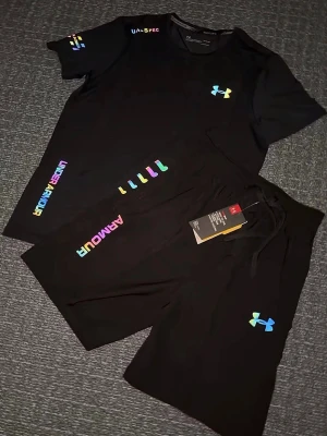 Svart träningsset från Under Armour - Säljer en svart träningsset från Under Armour med färgglada loggor och textdetaljer i regnbågsfärger. Shortsen har snörning i midjan och är tillverkade i ett mjukt och lätt material som passar perfekt för träning eller chill. Klassisk passform och sportig look. Storlek S, helt ny 