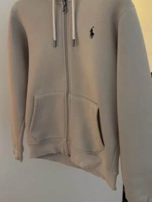 Beige hoodie med dragkedja från Polo Ralph Lauren - Snygg beige hoodie med dragkedja från Polo Ralph Lauren. Klassisk design med huva och vita dragsnören, två stora fickor framtill och den ikoniska loggan broderad på bröstet. Perfekt för en chill och stilren look.