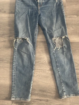 Blå mom jeans med slitna knän - Säljer ett par blå mom jeans med raka ben och stora slitningar över båda knäna. Jeansen har klassisk femficksmodell och är tillverkade i denim. Perfekta för en avslappnad och trendig look. Storlek 38, 32 längd 