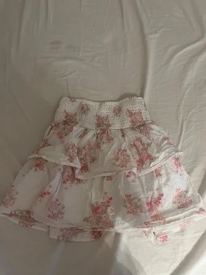 Vit blommig volangkjol - Söt kort kjol i vitt med rosa blommönster och bred smockresår i midjan. Kjolen har volangdetaljer och är gjord i ett lätt material som passar perfekt till varmare dagar. Romantisk och ungdomlig stil.