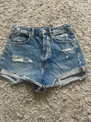 Blå högmidjade jeansshorts - Säljer ett par blå denimshorts med hög midja och slitna detaljer. Shortsen är uppvikta benslut och råa kanter för en avslappnad look. Perfekta för sommaren och ger en cool vintagekänsla. Storlek 36 men kan också passa till nån som är 32-34 