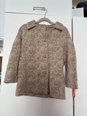 Beige mönstrad kappa med blommor - Snygg beige kofta med blommigt mönster och dekorativa guldknappar. Koftan har bred krage och rak passform, perfekt för dig som gillar vintagevibbar och unika detaljer. Materialet är tjockt och strukturerat, vilket ger en exklusiv känsla. Finns tillhörande band till🙏🏻