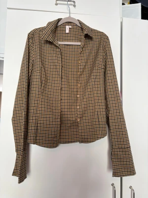 Brun och beige rutig skjorta - Snygg rutig skjorta i brunt och beige med klassisk krage och långa ärmar. Skjortan har knappar framtill och ett tidlöst rutmönster som passar perfekt till jeans eller kjol. Figursydd