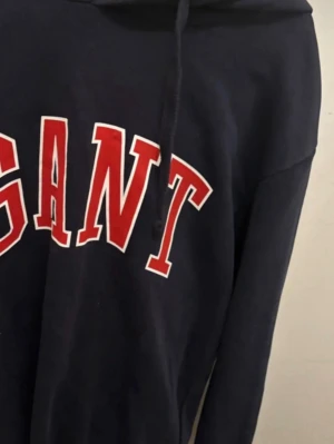 Mörkblå hoodie från GANT - Snygg mörkblå hoodie från GANT med stor röd och vit logga över bröstet. Klassisk huva med dragsko och mjukt material som känns skönt mot huden. Storlek L, men den känns mindre än en large