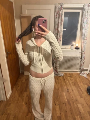 Beige stickad loungewear-set - Mysigt loungewear-set i beige stickat material med kabelstickat mönster. Setet består av en croppad hoodie med dragkedja och snörning samt matchande byxor med resår och dragsko i midjan. Perfekt för chill dagar hemma.