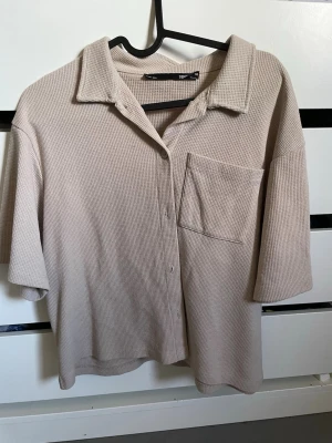 Beige ribbad skjorta med kort ärm - Säljer en beige skjorta i ribbat material med korta ärmar och en bröstficka. Skjortan har avslappnad passform, klassisk krage och knappar framtill. Perfekt för en chill och stilren look.