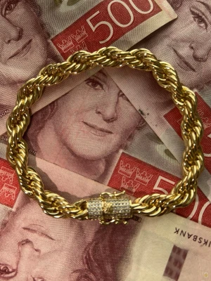 Guldpläterat Cordell Armband med Diamantlås - Säljer ett riktigt snyggt armband i guldfärgad metall med tvinnad design. Låset är dekorerat med små glittrande stenar som ger extra bling. Perfekt för dig som gillar statement-smycken och vill sticka ut lite extra.