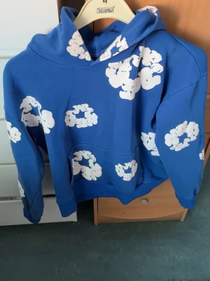 Denim tears hoodie  - Säljer en blå hoodie med stora vita molnliknande tryck över hela tröjan. Hoodien har en klassisk huva och magficka framtill. Perfekt för dig som vill sticka ut med en färgglad och lekfull stil. Materialet känns mjukt och bekvämt, perfekt för chill dagar.