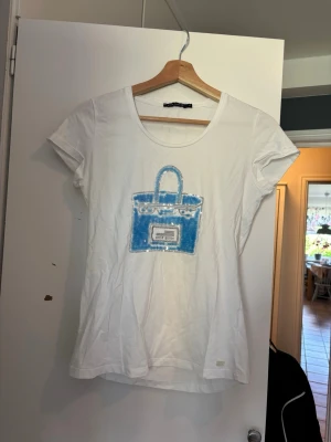 Vit t-shirt med blått tryck - Vit t-shirt i bomull med korta ärmar och ett stort blått tryck av en väska på bröstet. T-shirten har en klassisk passform och rund halsringning. Perfekt för dig som gillar enkla men ändå unika plagg. Den är i storlek M men passar bra på mig som har Xs/S