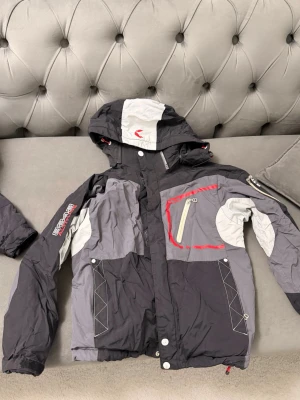 Svart och grå vindjacka med huva - vindjacka i svart och grått med röda detaljer och avtagbar huva. Jackan har flera fickor med dragkedja, justerbara muddar och coola kontrastsömmar. Sportig look med tryck på ärmen. Storlek 150. Pris går att diskutera 
