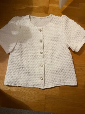 Vit strukturerad blus med pärlknappar - Säljer en vit blus med korta ärmar och rund halsringning. Blusen har en strukturerad, bubblig yta och fem dekorativa pärlknappar framtill. Passar perfekt till jeans eller kjol för en trendig look.