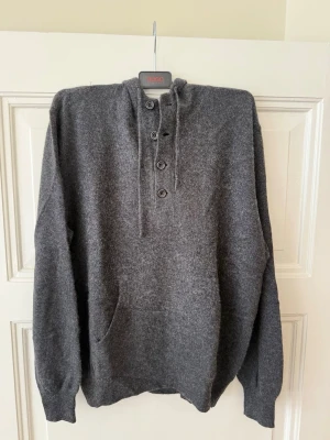 Grå cashmere hoodie från LINCS - Mysig grå stickad hoodie från LINCS i 100% cashmere. Tröjan har knappar framtill, huva och en stor magficka. Perfekt för dig som gillar stilrena och bekväma plagg med en lyxig känsla.