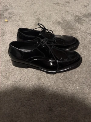 Svarta lackloafers med snörning - Säljer ett par svarta loafers från Viani i glansigt lackmaterial. Skorna har klassisk form med rundad tå och snörning framtill. Perfekta för dig som vill ha en stilren och trendig look. Passar till många olika outfits och ger en clean vibe. Pris kan diskuteras.