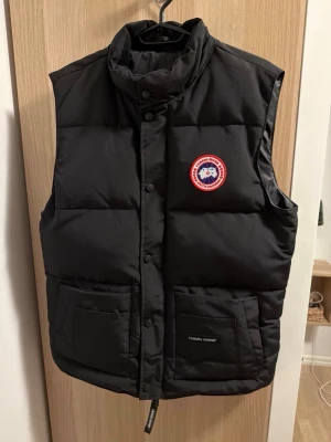 Svart dunväst från Canada Goose - Snygg svart dunväst från Canada Goose med klassisk logga på bröstet och två stora fickor framtill. Västen har hög krage och stängs med både dragkedja och tryckknappar. Perfekt för lager-på-lager och håller dig varm under kyliga dagar.