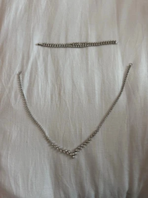 Festsmycken-set - Säljer ett elegant halsband i silverfärg med gnistrande strass-stenar i V-formad design. Setet levereras i sin eleganta originalask. Ett givet val för bröllop, bal, nyår eller för dig som älskar äldre festsmycken!Material: Silverfärgad metall (oädel metall/bijouteri) och infattade, fasetterade strasstenar med fin lyster.