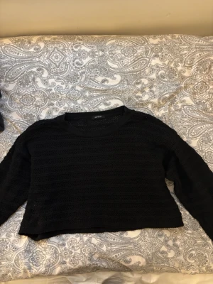 Svart virkad cropped tröja - Snygg svart stickad cropped tröja med virkat mönster och genombrutna detaljer. Tröjan har rund halsringning och långa ärmar. Perfekt att styla över ett linne eller t-shirt för en trendig lager-på-lager-look.
