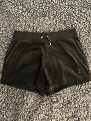 Svarta velourshorts Juicy Couture - Säljer ett par svarta velourshorts från Juicy Couture med snörning i midjan och fickor på sidorna. Klassisk logga broderad på ena benet. Perfekta för en chill och avslappnad stil.