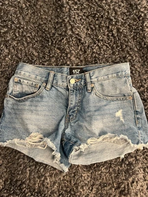 Blås denimshorts med slitningar - Säljer ett par blå denimshorts med råa, fransiga kanter och slitna detaljer framtill. Klassisk femficksmodell med knapp och dragkedja. Perfekta för sommaren och ger en avslappnad vibe.