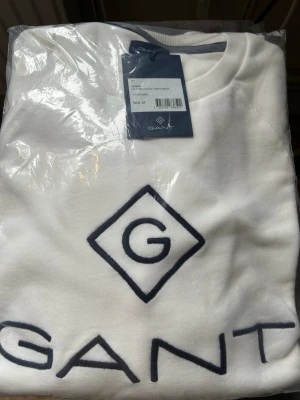 Vit sweatshirt från GANT - Säljer en vit sweatshirt från GANT med stor broderad logga och text på bröstet. Tröjan har rund halsringning och är tillverkad i mjukt material. Nypris 1049kr