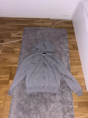 Grå hoodie från Polo Ralph Lauren - Klassisk grå hoodie med dragkedja från Polo Ralph Lauren. Tröjan har huva, långa ärmar och den ikoniska mörkblå loggan broderad på bröstet. Tillverkad i mjukt sweatshirtmaterial och har ribbade muddar vid ärmslut och nederkant.