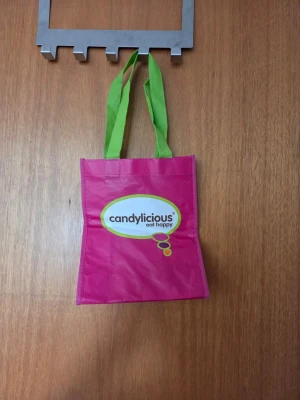 Rosa shoppingväska från Candylicious - Snygg rosa shoppingväska från Candylicious med gröna handtag och tryckt logga på framsidan. Väskan är tillverkad i slitstark plast och har en rektangulär form som rymmer mycket trotts liten. Perfekt för shoppingrundan eller att bära med sig småsaker.
