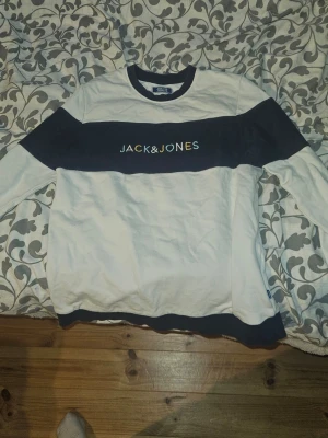 Vit och marinblå törja Jack & Jones - Snygg hoodie från Jack & Jones i vitt med breda marinblå paneler över bröstet, ärmarna och nederkanten. Stor logga framtill i kontrastfärg. Klassisk passform med rund hals och långa ärmar. Perfekt för en avslappnad streetwear-look.