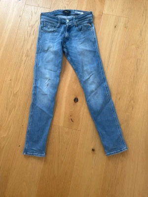 Blå slim fit anbass jeans från Replay - Snygga ljusblå skinny jeans från Replay med klassisk femficksdesign och dragkedja fram. Jeansen har slitningar och tvättade detaljer för en cool look. Perfekta för dig som gillar en smal passform och stilren design. Jeansen har en skada på baksidan!! Storleken är Waist: 28 och längd: 30 pris går att diskutera 