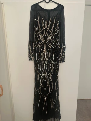 Svart långklänning med paljetter - Säljer en svart långklänning med glittriga paljettdetaljer i silver. Klänningen har lång ärm i transparent mesh och ett elegant mönster framtill och på ärmarna. Perfekt för dig som vill sticka ut med en glamorös look.