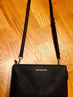 Svart axelväska från Michael Kors - Snygg svart axelväska från Michael Kors i ett tåligt, strukturerat material med silverfärgade detaljer och justerbar axelrem. Väskan har en stilren, rektangulär form och loggan i silver framtill.