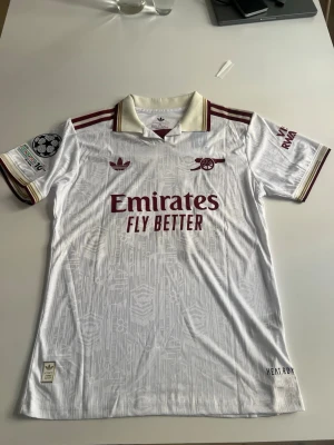 Helt ny! Arsenal 25/26 tredje Calafiori - Säljer en vit Arsenal matchtröja från Adidas med vinröda detaljer och klubbmärke. Tröjan har trycket 'Emirates Fly Better' på bröstet, Calafiori och nummer 33 på ryggen samt Champions League-märke på ärmen. Klassisk krage och korta ärmar. Perfekt för fotbollsfans!