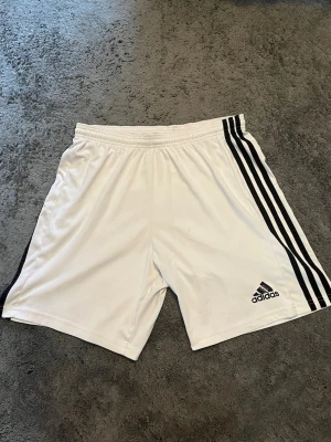 Vita Adidas shorts  - Snygga vita Adidas shorts med klassiska svarta ränder längs båda sidorna och Adidas-logga på vänster ben. Elastisk midja för skön passform, därmed perfekta för sport eller likande utövning. Tillverkade i lätt och bekvämt syntetmaterial. Pris går att diskutera!