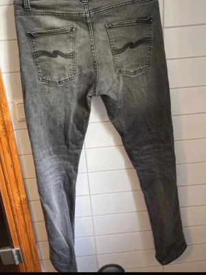 Grå lean dean jeans från Nudie Jeans - Säljer ett par grå lean dean jeans från Nudie Jeans med klassisk femficksmodell och svarta sömmar på bakfickorna. Jeansen har en snygg tvättad look och är tillverkade i bomull med stretch för extra komfort.
