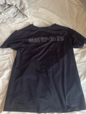 Svart tränings-tshirt från MM Sports - Säljer en svart tränings-tshirt från MM Sports i storlek M. T-shirten har rund halsringning och tryckt logga på både bröstet och ryggen. Tillverkad i ett lätt och snabbtorkande material som passar perfekt till gymmet eller löpning.