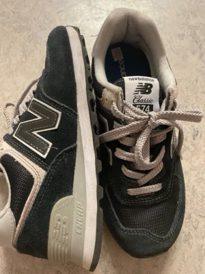 New Balance 574 svarta sneakers - Klassiska New Balance 574 sneakers i svart mocka och mesh med vita detaljer och grå sula. Skorna har den ikoniska N-loggan på sidan och vita skosnören. Perfekt för dig som gillar retrostil och bekväma sneakers.