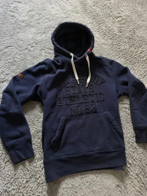 Mörkblå hoodie från Superdry - Snygg mörkblå hoodie från Superdry med broderad text på bröstet och stor känguruficka framtill. Hoodien har vita dragsnören i huvan och en liten patch på ärmen. Tillverkad i mjukt sweatshirtmaterial, perfekt för en avslappnad stil.