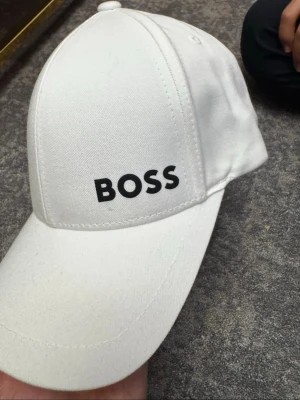 Vit keps från BOSS - Snygg vit keps från BOSS med klassisk böjd skärm och svart BOSS-logga framtill. Tillverkad i bomullstyg med justerbar rem bak för perfekt passform. Enkel och stilren design som passar till det mesta.