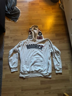 hoodie från Hoodrich - Säljer en vit hoodie från Hoodrich med stor svart logga på bröstet och textdetaljer på huvan och ryggen. Hoodien har känguruficka framtill och ribbade muddar. Perfekt för dig som gillar streetwear och vill sticka ut med coola detaljer.