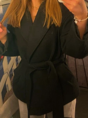 Svart kappa med knytskärp - Stilren svart kappa med bred krage och knytskärp i midjan. Modellen är rak och har en avslappnad passform, perfekt för lager på lager. Kappan har långa ärmar och är tillverkad i ett mjukt tyg som ger en snygg siluett. Kappan är nopprig därav priset. 