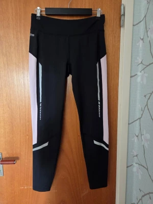 Svarta leggings med rosa detaljer - Snygga svarta leggings med rosa och silvriga detaljer längs sidorna och benen. Hög midja och stretchigt material som sitter skönt på kroppen. Perfekta för träning eller chill dagar. Reflekterande detaljer för extra stil.