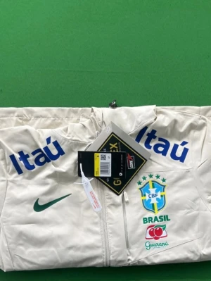 Vit Brasil Nike träningsjacka - Snygg vit träningsjacka från Nike med Brasiliens landslagslogga och flera sponsortryck i blått och grönt. Jackan har hel dragkedja, hög krage och tryckta detaljer på bröst och ärmar. Perfekt för dig som älskar fotboll och vill ha en unik look.