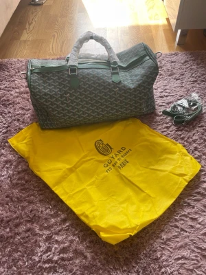 Grön weekendbag - Snygg grön weekendbag från med klassiskt mönster och detaljer i grönt läder. Väskan har dubbla handtag, avtagbar axelrem och kommer med en gul dustbag. Perfekt för resor när du vill ha både stil och funktion.