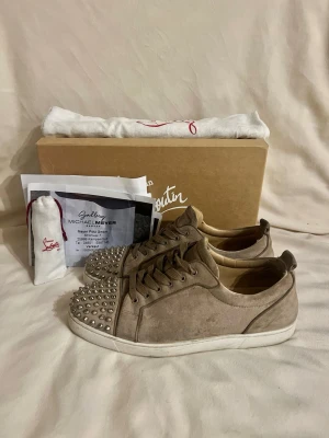 Christian Louboutin - Säljer ett par beige sneakers från Christian Louboutin i mocka med silverfärgade nitar på tån. Skorna har klassisk låg profil, vita sulor och ton-i-ton snörning. Perfekta för dig som vill sticka ut med exklusiv design och edgy detaljer. Efter en liten tvätt är de perfekta!