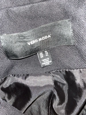 Svart jacka/rock/ coats från Vero Moda - Svart jacka/ rock /coats  från Vero Moda med klassisk rak passform och två knappar framtill. Rocken har ett slätt, glansigt innerfoder och är sydd i ett tyg med diskret struktur. Perfekt för dig som gillar en stilren och enkel look.