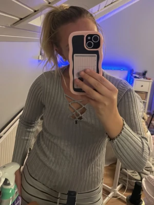 Ljusgrå ribbad långärmad topp - Säljer en ljusgrå ribbad topp med lång ärm och snygg snörning framtill i halsringningen. Toppen har en figurnära passform och är perfekt för dig som gillar stilrena detaljer.