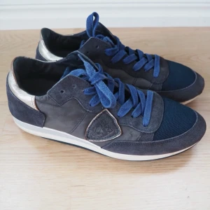 Philippe model mörkblå sneakers med metallic-detalj - Sonen har vuxit ur sina fina Philippe Model skor. Snygga mörkblå sneakers med blå skosnören, mesh framtill och detaljer i mocka och läder. Skon har en metallic guldaktig häl och vit sula med blå undersida. Perfekt för dig som gillar en sportig och trendig look.                                                                   Innermått 24,5cm.