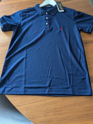 Marinblå pikétröja från Ralph Lauren - Klassisk marinblå pikétröja från Ralph Lauren med korta ärmar, krage och tre knappar framtill. Tröjan har den ikoniska röda logotypen broderad på bröstet. Perfekt för en stilren och avslappnad look.