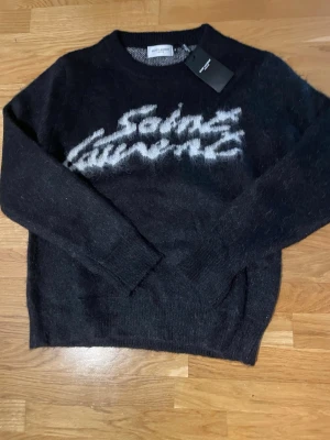 Svart Saint Laurent stickad tröja - Säljer en svart stickad tröja från Saint Laurent med vit logotyptext framtill. Tröjan har rund halsringning, långa ärmar och är gjord i ett fluffigt, mjukt material som ger en lyxig känsla. Perfekt för dig som vill ha något trendigt och bekvämt.